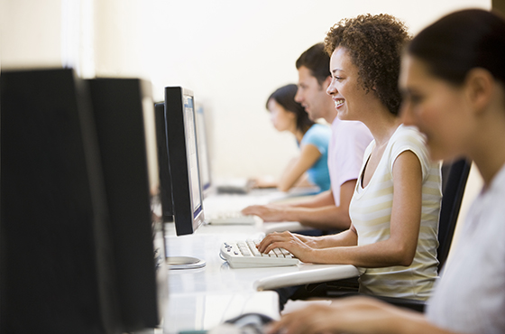 Four students sitting at a long table working on desktop computers